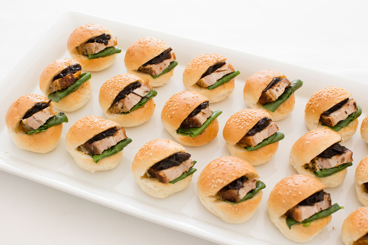 Mini pork sliders with spinach and dark sauce on a white serving platter, showcasing a delicious and elegant catering option.