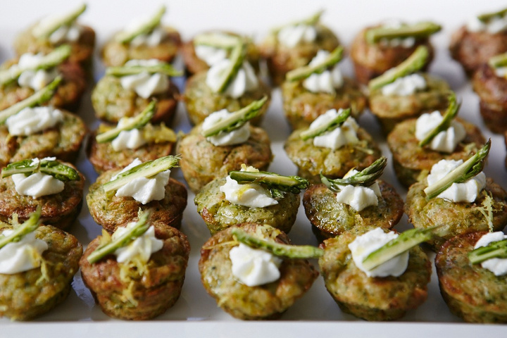 Close-up of savory muffins on a white platter, each topped with white cream and a sprig of asparagus, showcasing elegant finger food.