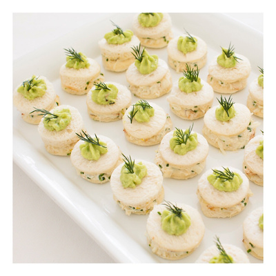 Close-up of a platter of round appetizers, each topped with green sauce and a sprig of dill, on a white background.