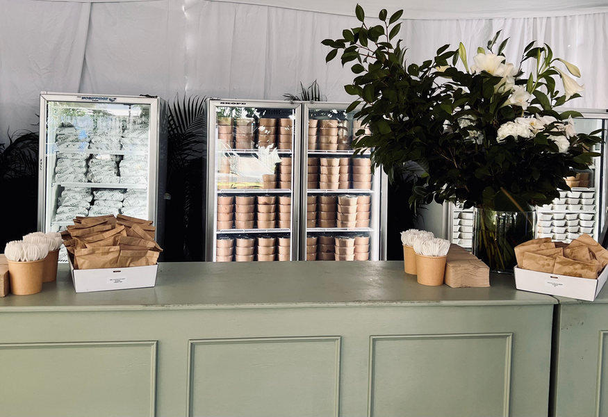 Refrigerated display cases behind a counter featuring white flower arrangements, brown paper bags, and paper cups at an indoor event.