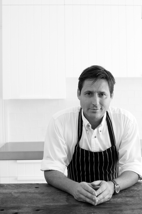 Black and white portrait of an adult male chef in a striped apron with hands clasped, set against a minimalist kitchen backdrop.