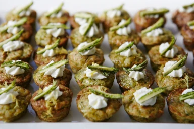 An overhead view of savory mini tarts topped with cream cheese and asparagus spears arranged neatly on a white tray.