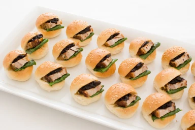 An array of miniature sliders with roasted pork and greens arranged neatly on a long white rectangular serving platter.