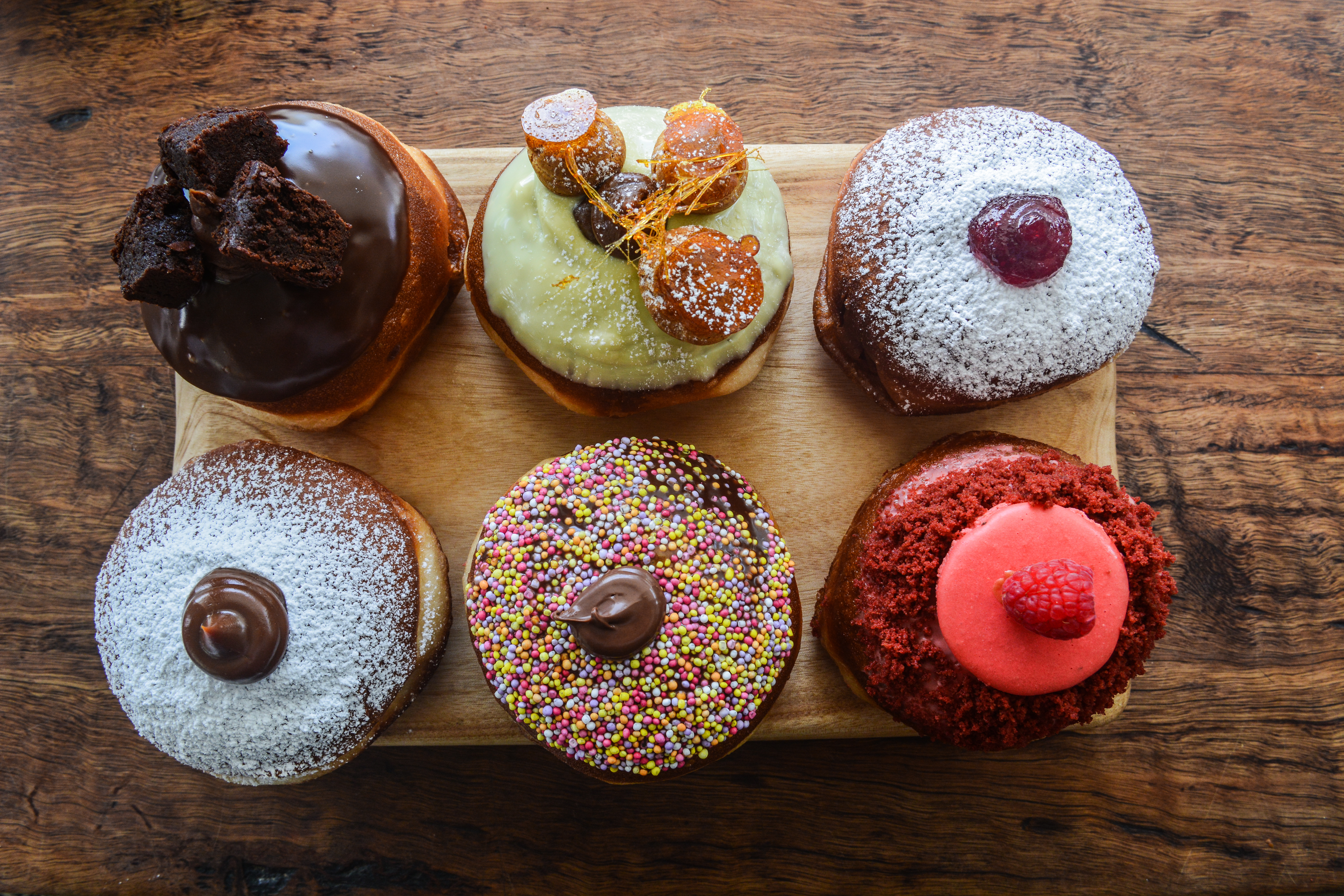 Overhead shot of six decorated donuts on a wooden cutting board, featuring chocolate, sprinkles, icing, and assorted toppings. Food photography.