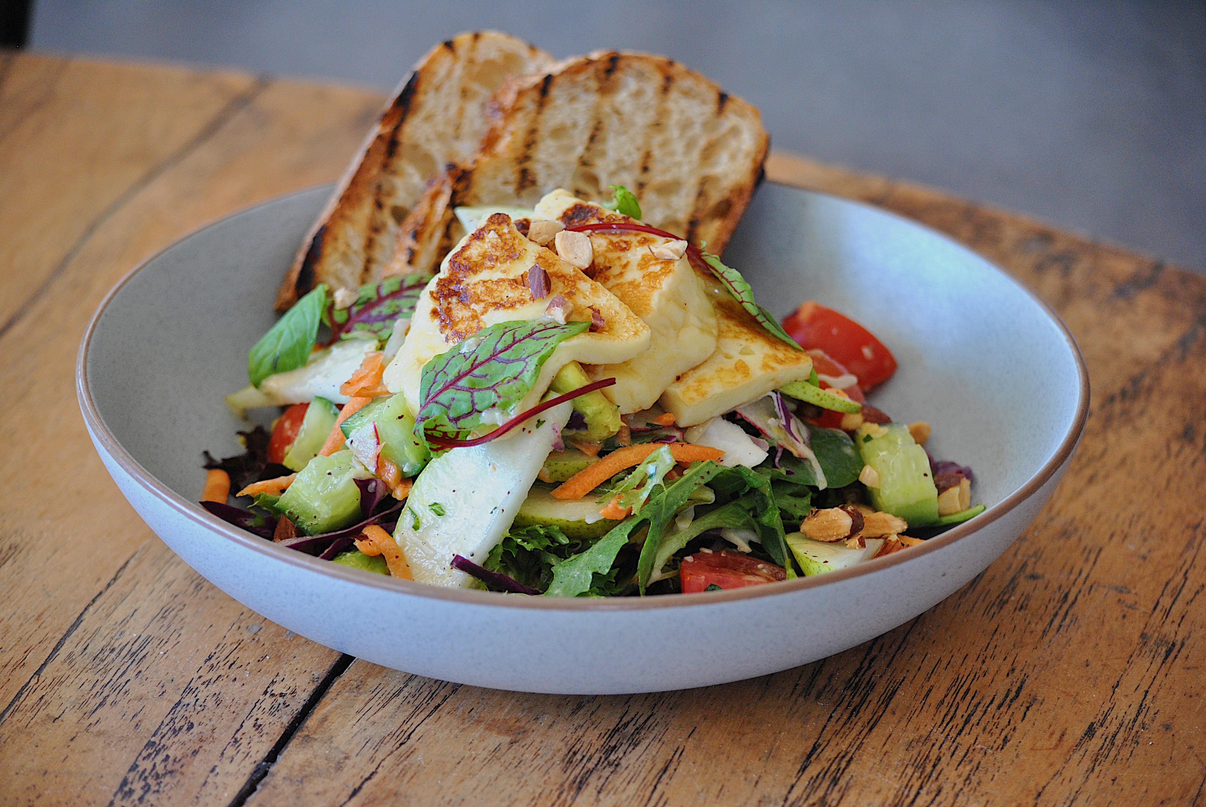Close-up of a halloumi salad in a bowl on a wooden table, featuring grilled halloumi cheese, sliced vegetables, greens, and grilled bread.