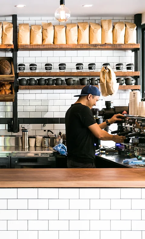 Barista prepares espresso at a coffee shop, brown paper bags with coffee beans on a shelf, white subway tile, and a dark wood counter.