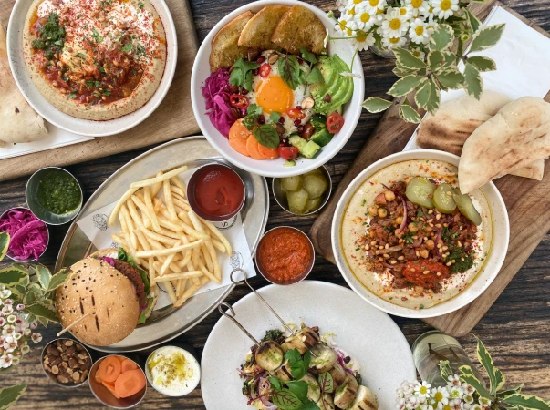 Overhead shot of a wooden table laden with Middle Eastern cuisine including hummus, a grain bowl, a burger, and grilled vegetable skewers.