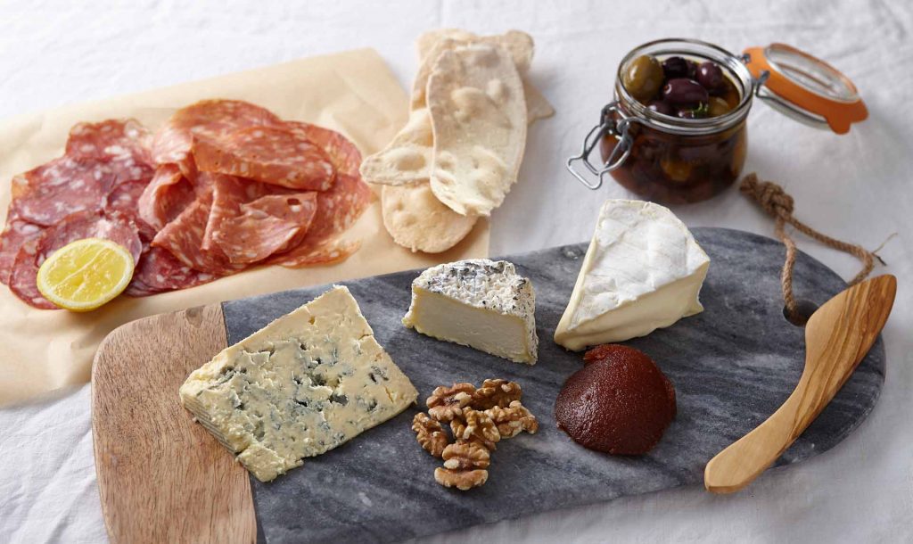 Overhead shot of charcuterie board with salami, cheese, crackers, walnuts, quince paste, and olives in a jar on white background.
