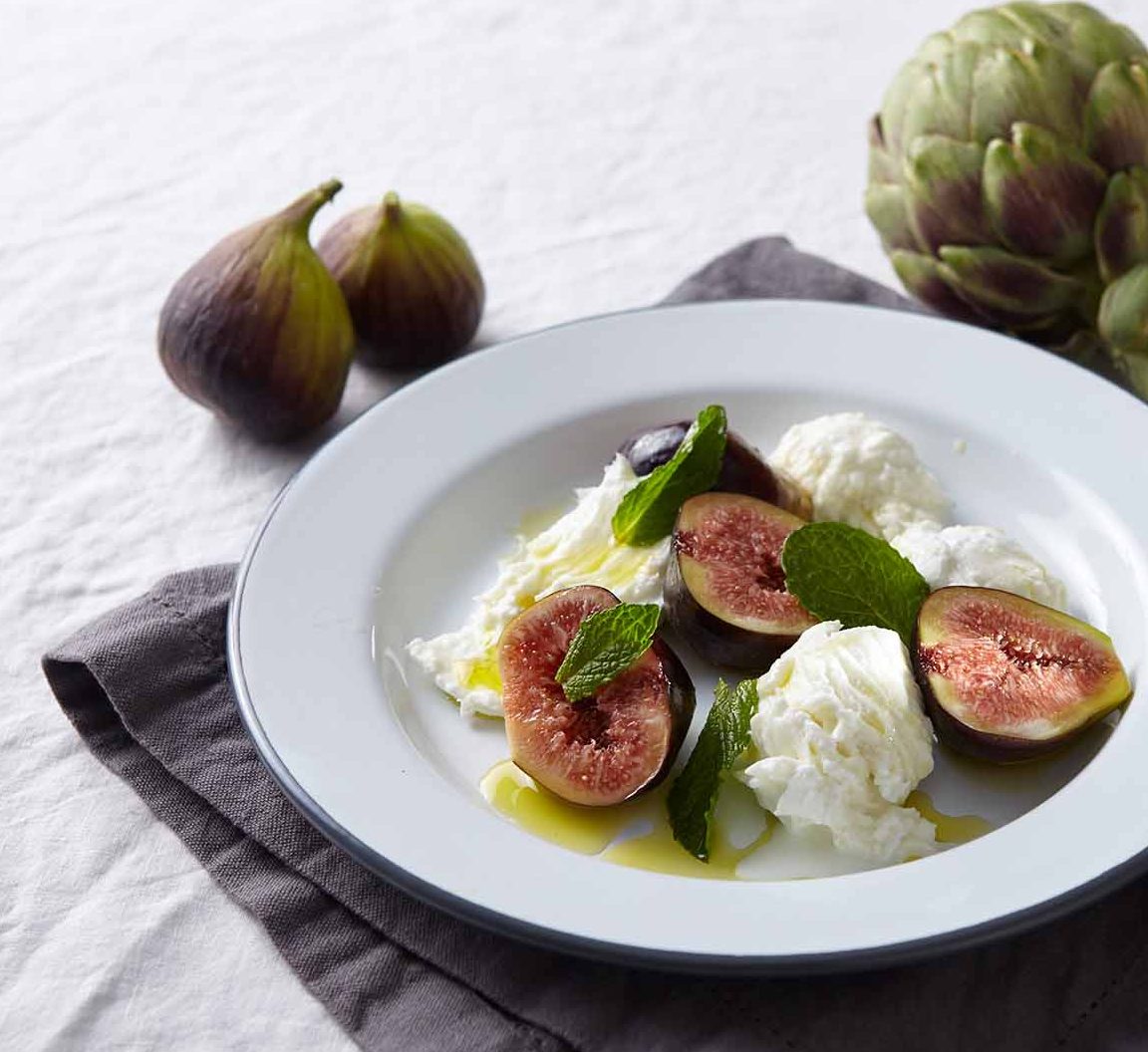 Overhead shot of a white plate with sliced figs, ricotta cheese, mint leaves, and artichokes; food photography, studio lighting.