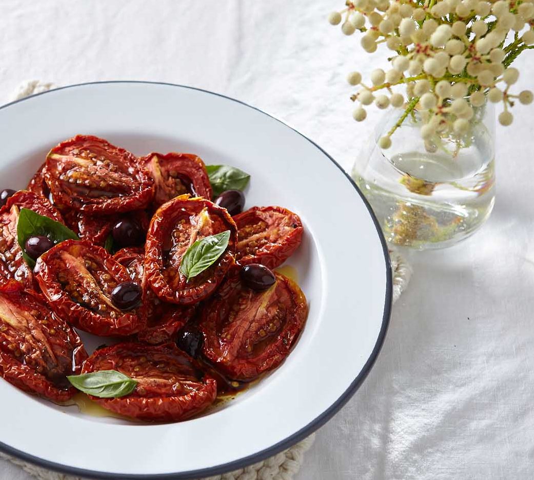Close-up of a white plate with sun-dried tomatoes, basil leaves, and olives, set against a white linen background with a flower vase.