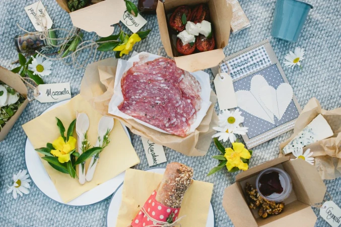 Overhead picnic blanket scene with cured meats, cheese, bread, and boxed appetizers arranged with fresh flowers and name tags for food items.
