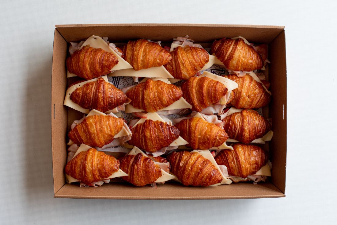 Overhead shot of a brown cardboard box filled with croissants, cheese and ham sandwiches on a white surface, food photography.