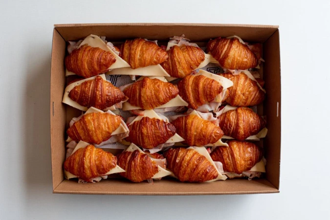 Overhead shot of a brown cardboard box filled with croissants, cheese and ham sandwiches on a white surface, food photography.