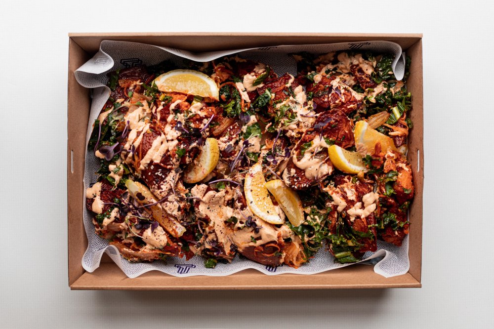 Overhead shot of a box filled with pieces of food, topped with sauce, greens, and lemon slices, against a white background.