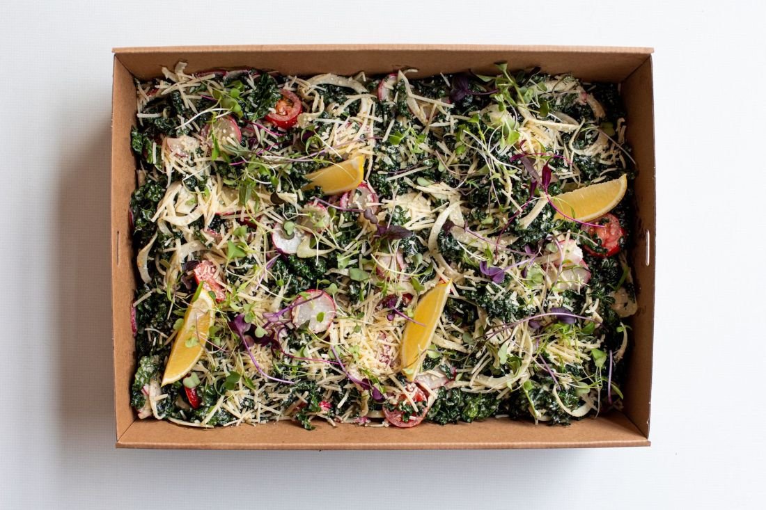 Overhead shot of a cardboard tray with a salad featuring kale, white shredded cheese, sprouts, sliced radishes, and lemon wedges.