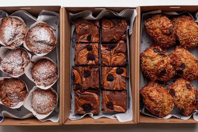 Three cardboard boxes filled with treats: muffins with powdered sugar, brownies with swirl patterns and toppings, and coconut muffins with chocolate chips.