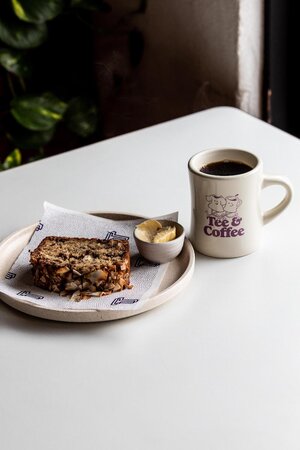 Overhead shot of a plate with a slice of banana bread, a small dish of banana slices, and a mug of coffee on a white table.