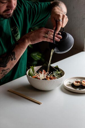 Person pouring liquid from a teapot into a white bowl filled with food on a white table, with chopsticks and a plate of eggs.