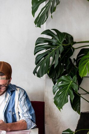 Adult male in striped shirt at table with tropical Monstera plant and white textured wall in a restaurant setting.