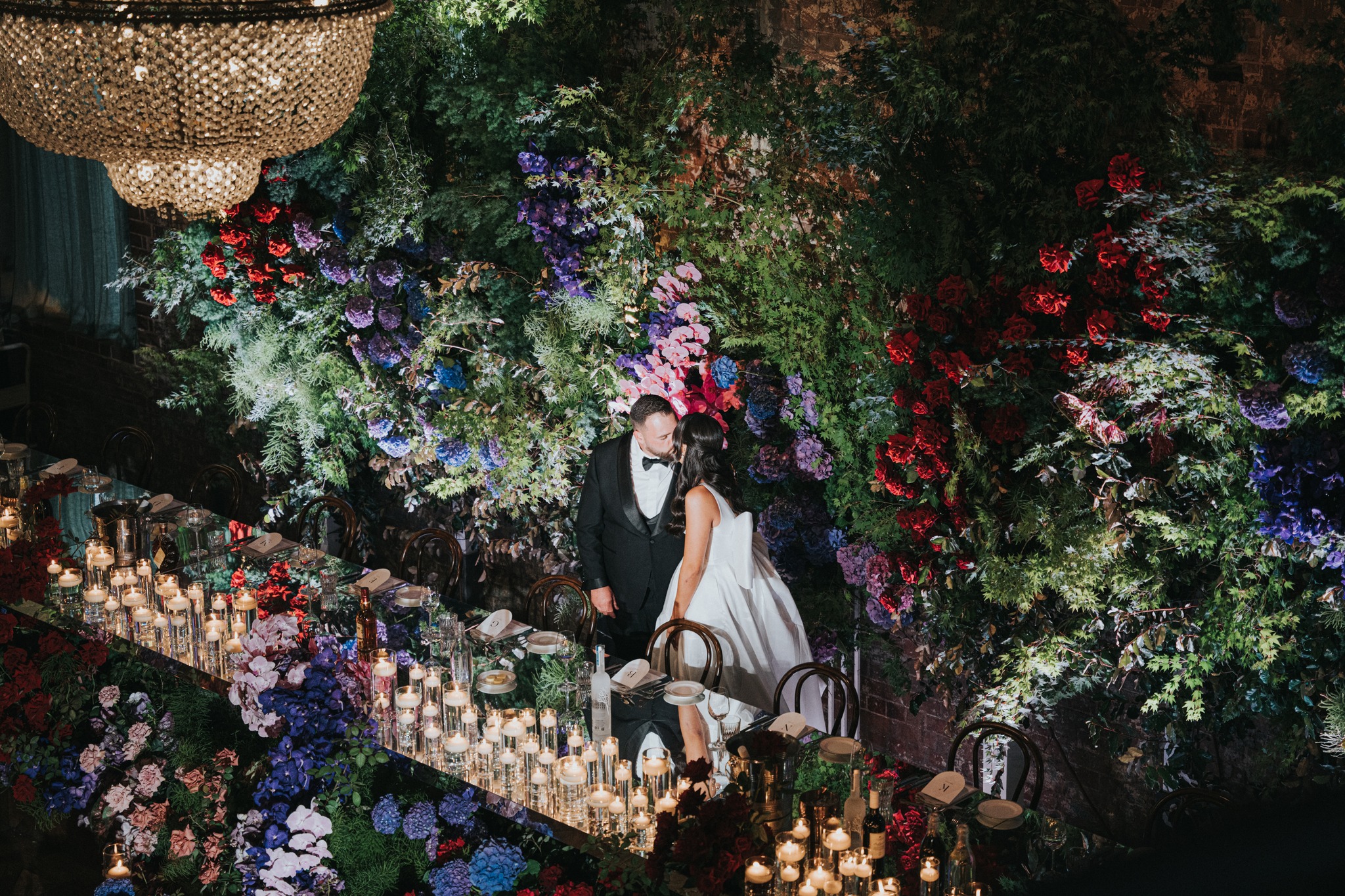 Wedding reception scene shows a bride and groom near a long table with flower arrangements, many candles, and a chandelier.