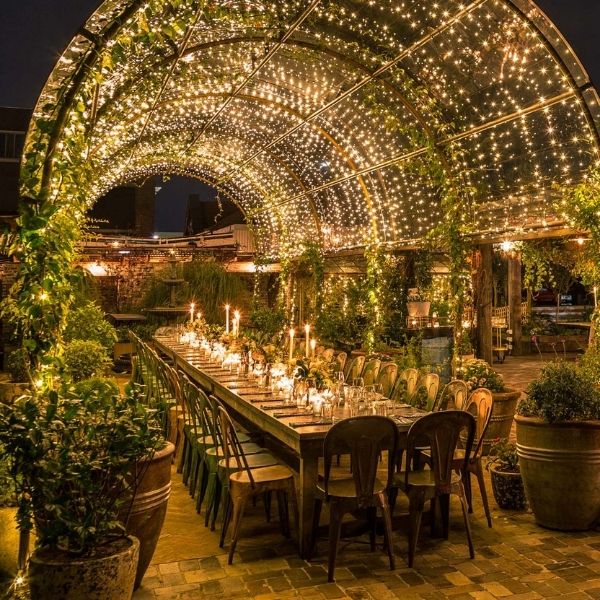 Outdoor dining area at night with a long table set for a gathering, illuminated by overhead string lights and surrounded by greenery.