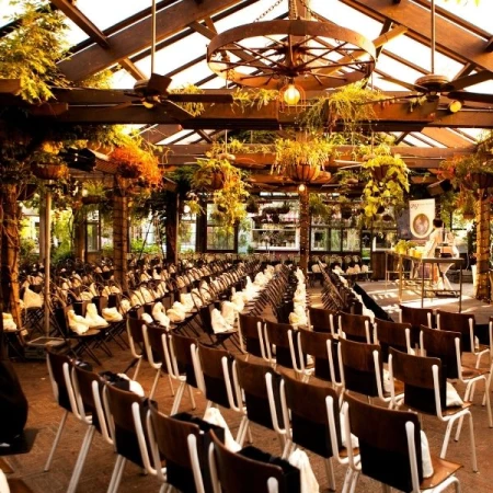 Indoor event with rows of chairs beneath plants hanging from the ceiling and rustic chandeliers, creating a warm and inviting atmosphere.