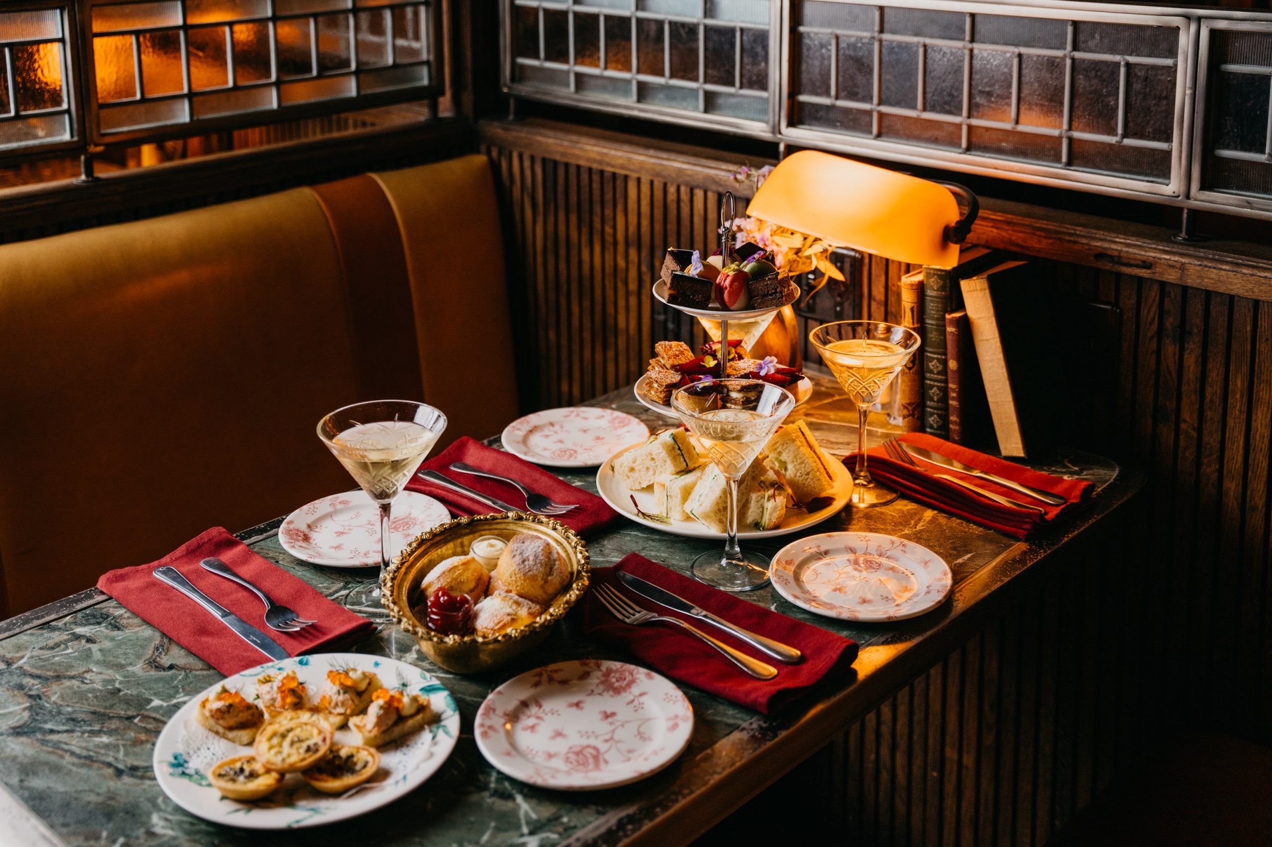 Elegant afternoon tea on a marble table with tiered desserts, finger sandwiches, scones, and cocktails in a warmly lit indoor setting.