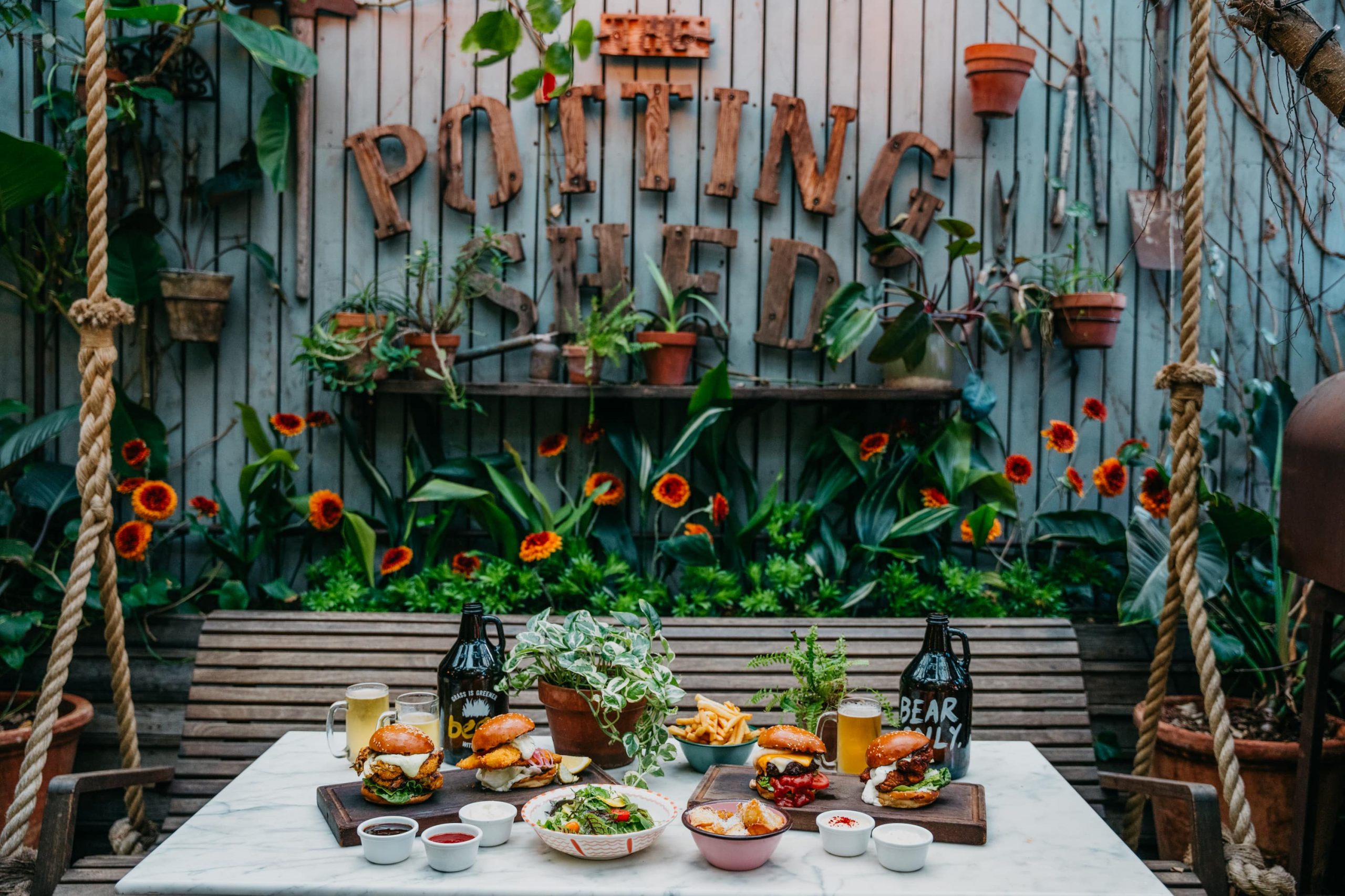 Outdoor dining table set for burgers and drinks at The Potting Shed restaurant, surrounded by green plants and rustic decor.