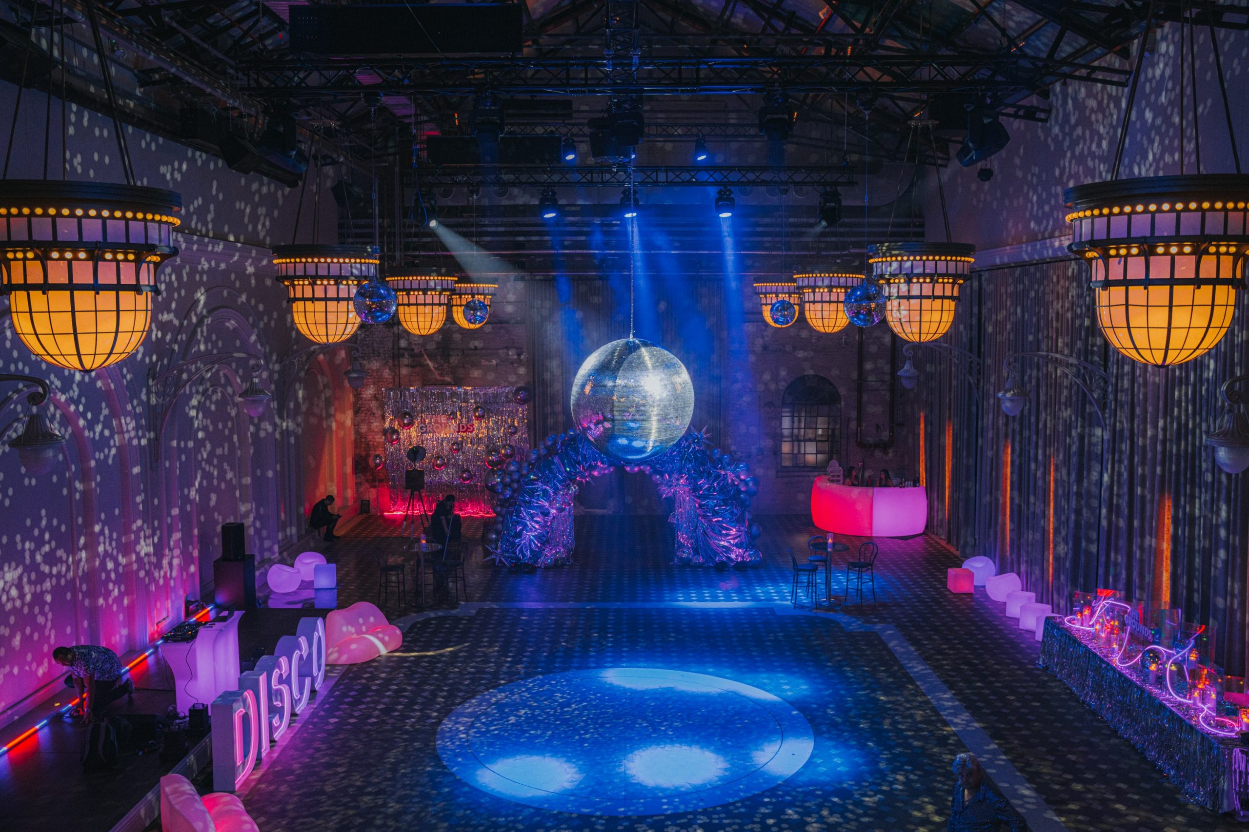Large room is decorated for a disco-themed event with hanging mirror balls, party lights, archway, and retro DISCO signage.