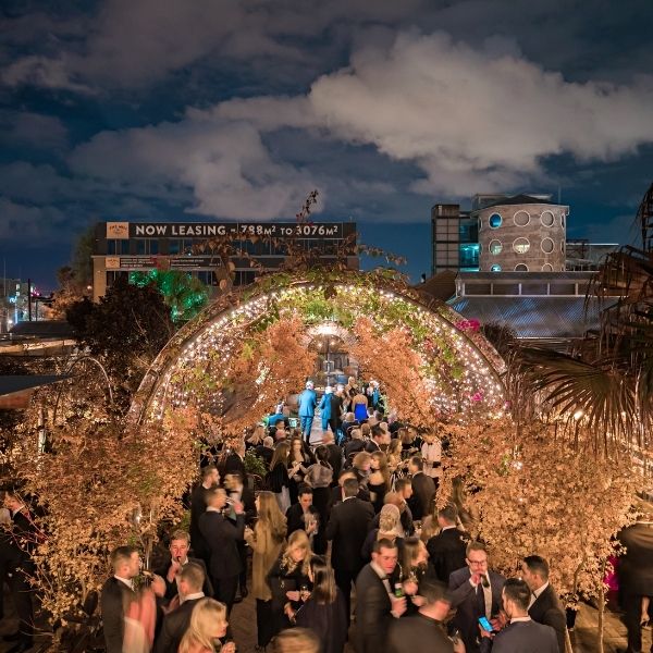 Large group of formally dressed people gather at an outdoor evening event, walking through a tunnel decorated with lights and greenery.