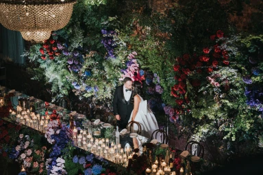 Bride in white gown and groom in black tuxedo kissing before a lush floral wall and candlelit table in Sydney.