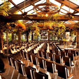 Rows of wooden chairs with white covers set up for an event under a rustic glass and timber pavilion adorned with hanging plants.