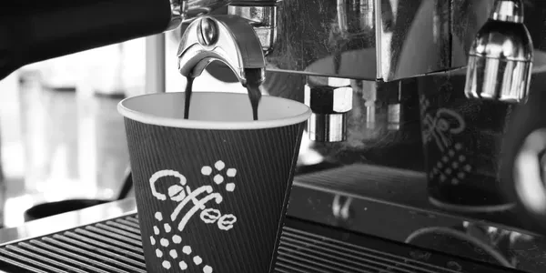 Close-up of a paper coffee cup being filled by a commercial espresso machine in a monochromatic palette, with a focus on textures.