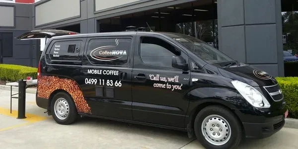 Black mobile coffee van parked outdoors with coffee bean design, company logo, and contact information displayed on the vehicle.