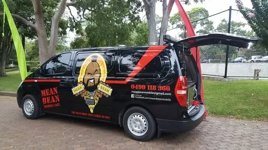 Black van with red and yellow graphics parked outdoors with an open back, promoting 'Mean Bean Mobile Cafe' on a paved surface.