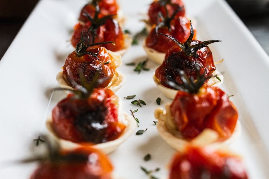 Close-up of a white plate with a row of roasted tomato appetizers in small pastry shells, garnished with fresh herbs.
