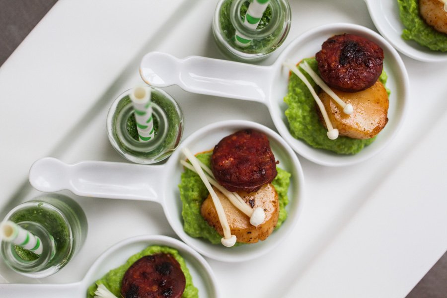 Appetizers on a white tray, including bite-sized chorizo and scallops, plus green smoothies with striped straws, overhead angle.