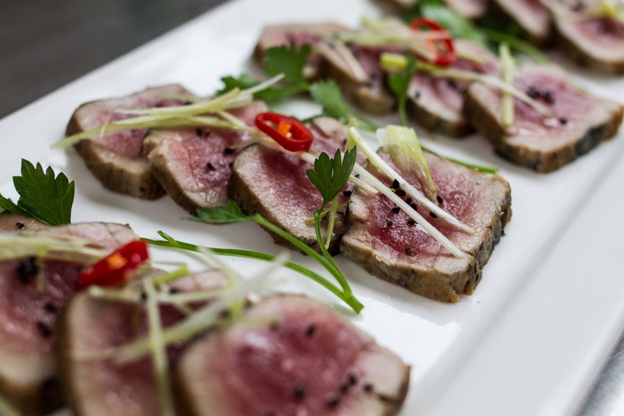 Seared tuna slices are arranged on a white plate, garnished with herbs, white onion, and red pepper; food photography.