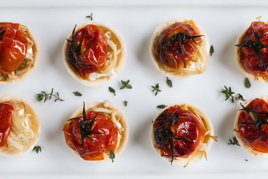 Overhead view of eight tartlets with roasted cherry tomatoes, cheese, and thyme arranged on a white rectangular surface.