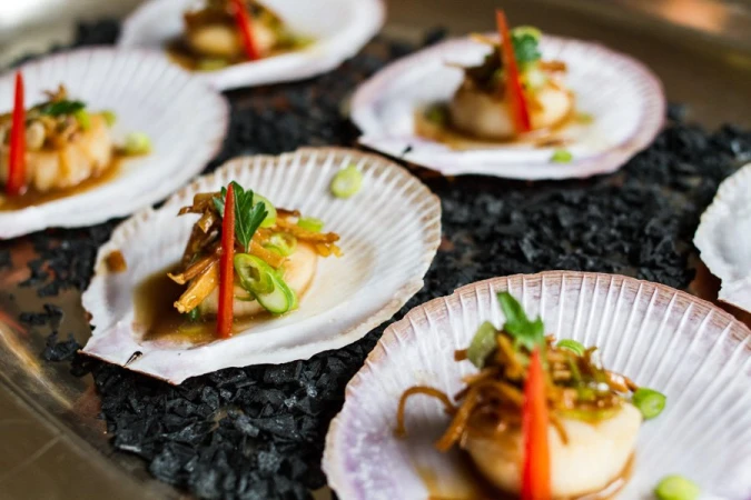 Seared scallops served in scallop shells on a bed of black pebbles with garnishes of herbs and sliced scallions.
