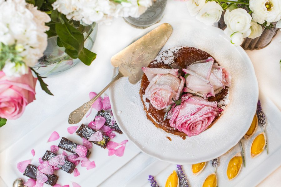 Overhead shot of a cake on a white plate, adorned with pink roses and powdered sugar, alongside chocolate bars and flower petals on a white surface.
