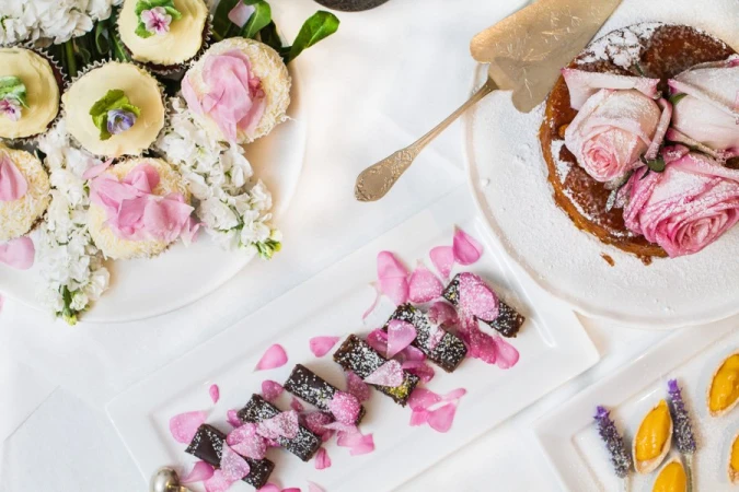 Overhead shot of dessert plates with cupcakes, chocolate bars, and a cake with flowers on a white tablecloth background.