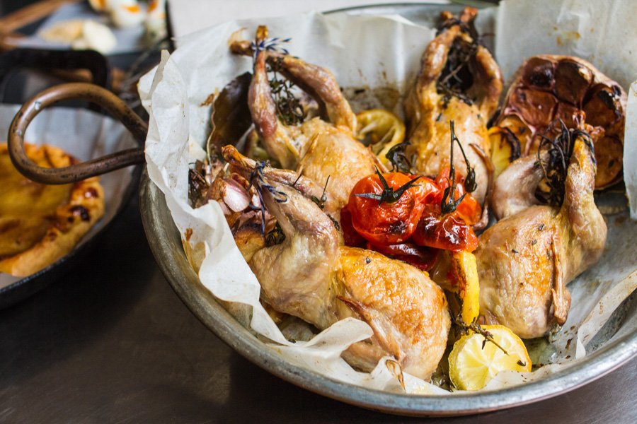 Roasted quail in a metal serving dish, accompanied by roasted tomatoes, garlic, lemon wedges, and herbs. Culinary presentation.