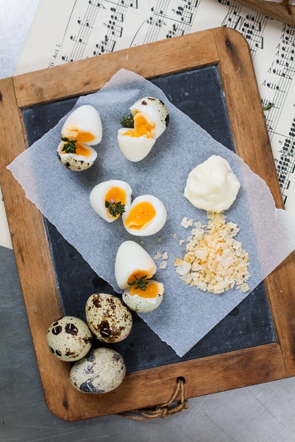Overhead shot of halved quail eggs with herbs on a wooden board, accompanied by a small amount of white sauce and seasonings.