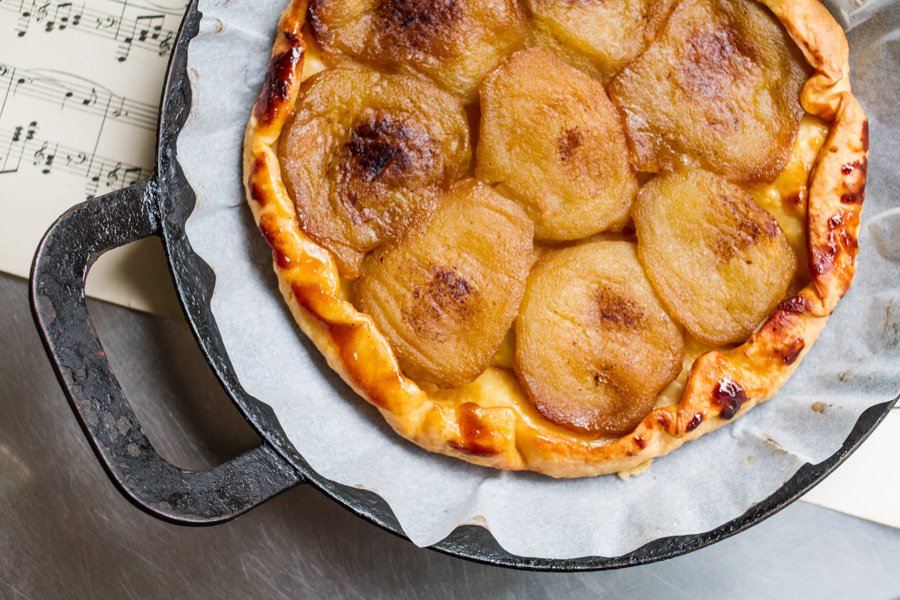 Overhead shot of a golden-brown pear tart with flaky crust in a black cast-iron pan, accented by a musical score backdrop.