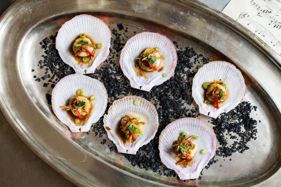 Overhead shot of six scallop dishes arranged on a silver platter with black granular accents, offering a fresh culinary perspective.