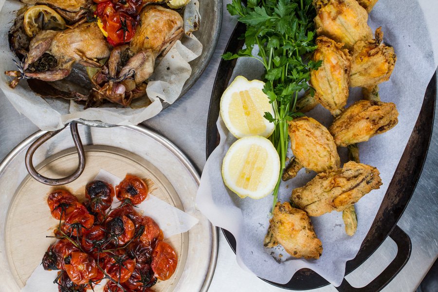 Overhead shot of three metal platters of roasted squab, tomatoes, and fried squash blossoms, accompanied by lemons and parsley.
