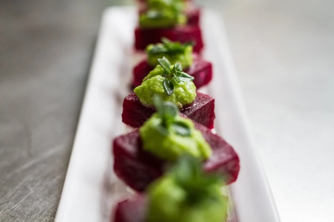 A row of vibrant appetizers on a long white plate featuring cubed beets topped with green puree and herb sprigs, creating a food presentation.