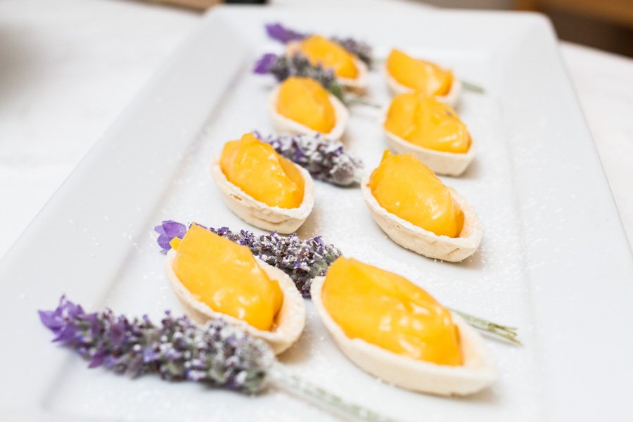 Overhead shot of an array of dessert tarts with a bright yellow filling, accented with lavender flowers on a white platter.