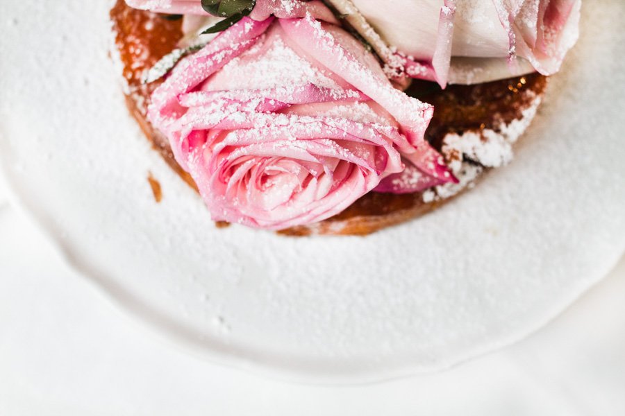 Overhead shot of dessert on a white plate. The baked good is topped with pink roses and powdered sugar, set on a white table cloth.